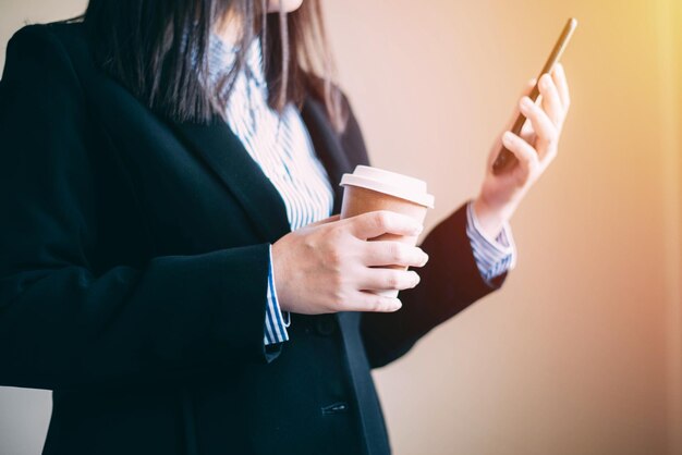 Środkowa sekcja bizneswoman trzymająca jednorazowy kubek podczas korzystania z telefonu komórkowego