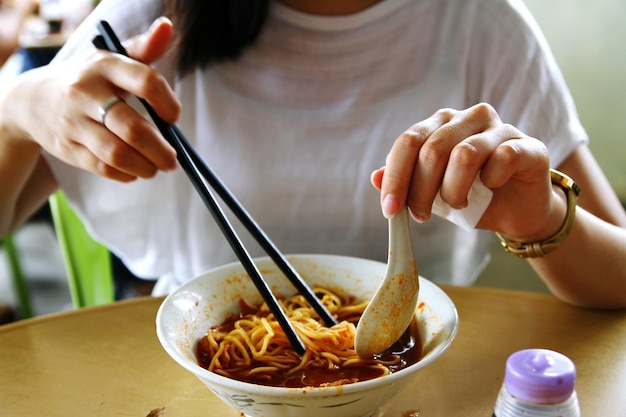 Środkowa sekcja kobiety trzymającej pałeczki i łyżkę nad miską hokkien mee