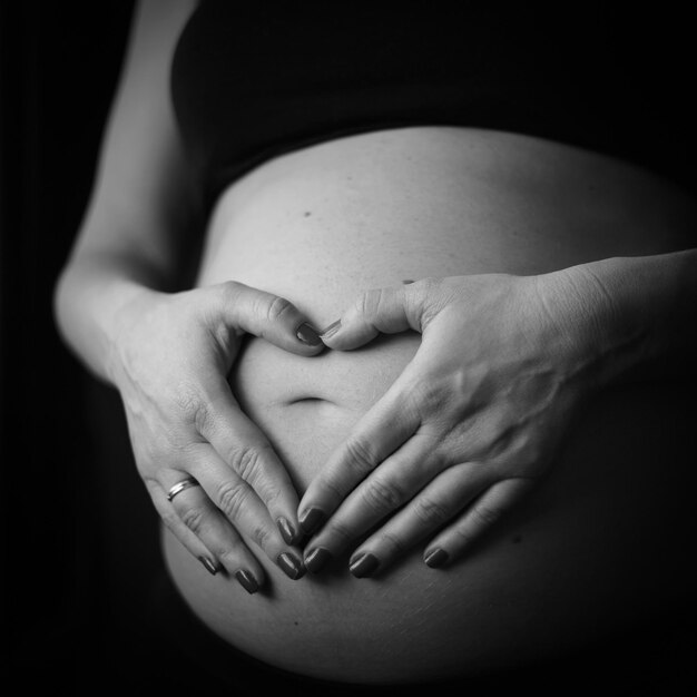 Zdjęcie Środkowy odcinek kobiety w ciąży w kształcie serca z rękami na brzuchu na czarnym tle