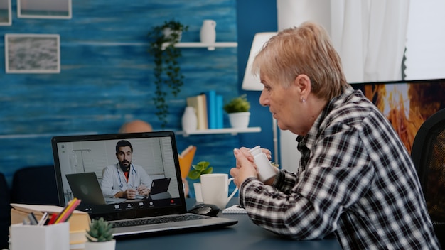 Stara kobieta siedzi przy stole w domu, patrząc na laptopa, odpowiadając na pytanie o zdrowie, rozmawia z terapeutą przez aplikację do wideokonferencji, trzymając butelkę z tabletkami Koncepcja zdalnej konsultacji medycznej