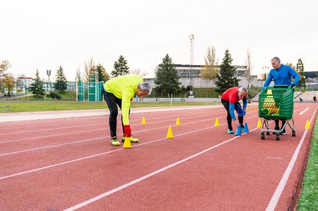 Zdjęcie starsi sportowcy na torze sportowym jeden z wózkiem z stożkami inny zgięcie, aby umieścić stożki