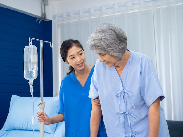 Starsza Azjatycka kobieta pacjentka próbuje chodzić po balkoniku trzymana i ostrożnie wspierana w ramionach przez opiekuna młoda uprzejma asystentka pielęgniarki w niebieskim garniturze w białym pokoju koncepcja opieki seniora