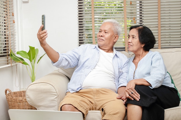 Starsza Azjatycka Para Ogląda Multimedia Online Na Swoim Smartfonie W Salonie W Domu. Koncepcja życia Po Przejściu Na Emeryturę