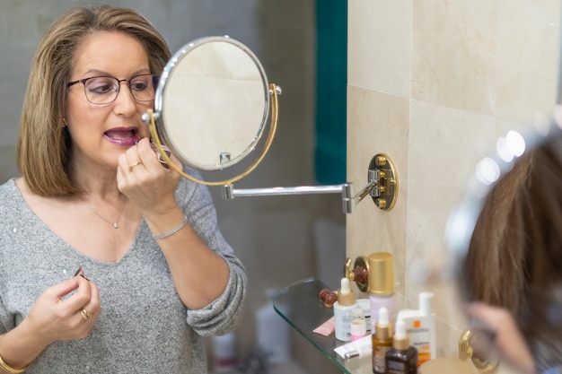 Starsza biała kobieta maluje usta przed lustrem w łazience