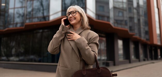 Starsza bizneswoman w stylowym płaszczu uśmiecha się podczas rozmowy na telefonie komórkowym poza nowoczesnym
