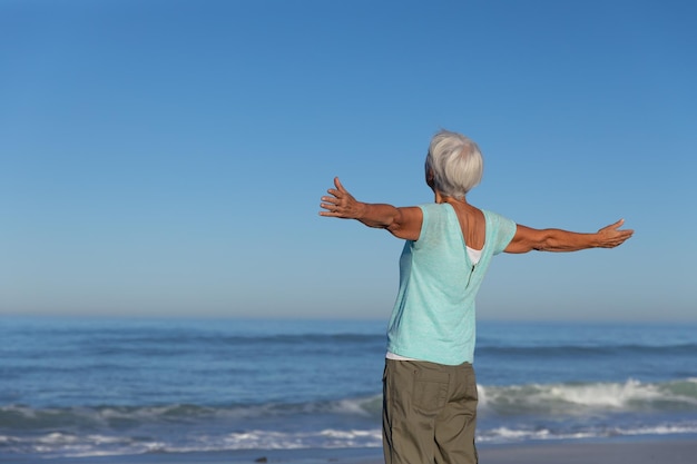 Starsza kaukaska kobieta ciesząca się czasem na plaży w słoneczny dzień, stojąca z wyciągniętymi ramionami i patrząca na morze, z błękitnym niebem w tle