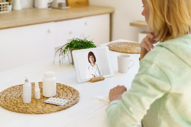 Zdjęcie starsza kobieta bez twarzy rozmawiająca online z lekarzem siedzącym w kuchni w domu