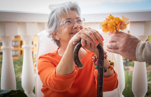 Starsza Kobieta Cierpiąca Na Ból Pleców Siedzi Z Rękoma Opierając Się Na Patyku, Gdy Odbiera Kwiatek Od Męża