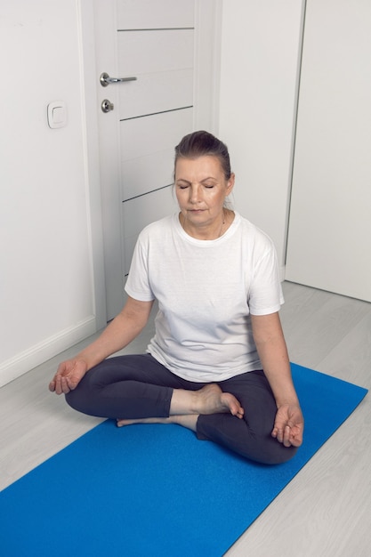 Starsza kobieta ćwiczy jogę na niebieskim dywanie w białym pokoju w domu