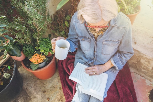 Starsza kobieta czyta książkę w otoczeniu roślin z kubkiem w dłoni