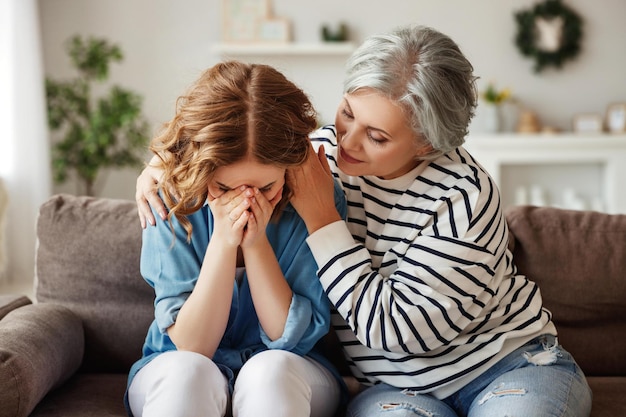 Starsza kobieta obejmuje i wspiera płacz córeczki siedząc razem na kanapie w domu