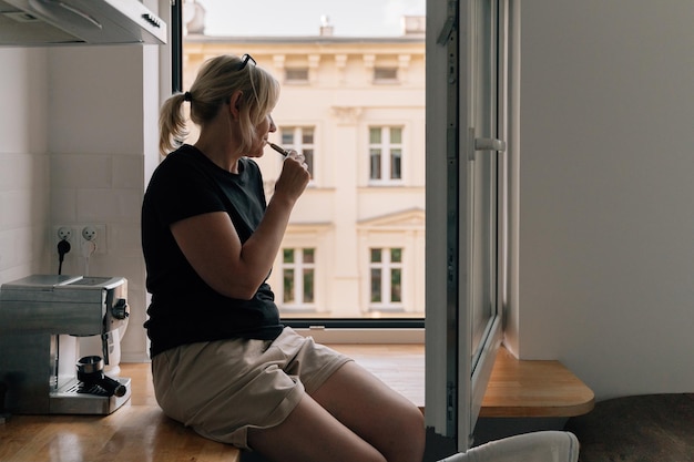 Starsza kobieta palenia e-papierosa w domu przy oknie Kaukaski 40 50-letnia kobieta palacz