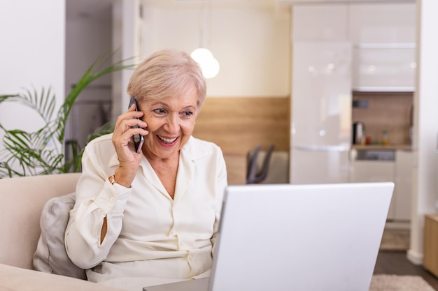 Starsza kobieta pracująca na laptopie, uśmiechająca się, rozmawiająca przez telefon, starsza kobieta korzystająca z laptopa, starsza żona siedząca w domu, korzystająca z laptopu i rozmawiająca na telefonie komórkowym, uśmiechnięta