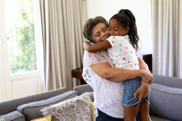 Starsza kobieta rasy mieszanej i jej wnuczka cieszą się razem w domu, stojąc przy kanapie, kobieta trzyma dziewczynę, obejmując i uśmiechając się