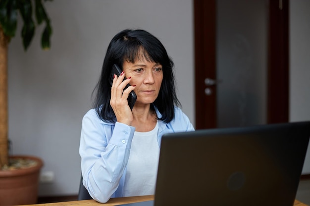 Starsza kobieta rozmawia przez telefon w swoim miejscu pracy z laptopem