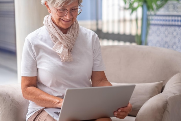 Starsza kobieta siedzi na kanapie podczas korzystania z laptopa Starsza atrakcyjna kobieta korzystająca z technologii i kontaktów towarzyskich