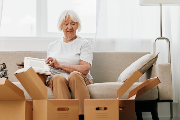 Starsza kobieta siedzi na kanapie w domu z pudełkami zbierającymi rzeczy z albumami wspomnień ze zdjęciami i ramkami do zdjęć przeprowadzającymi się do nowego miejsca, sprzątającymi i szczęśliwym uśmiechem Emerytura ze stylu życia