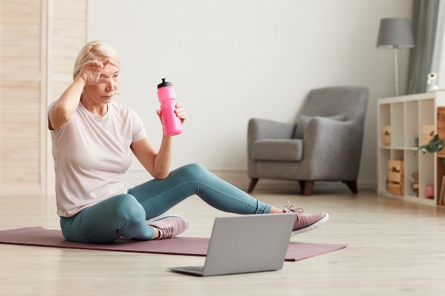 Starsza kobieta siedzi na podłodze na macie do ćwiczeń i wody pitnej po treningu sportowym w domu