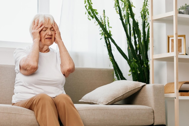 Starsza kobieta silny ból głowy siedząca na kanapie problemy zdrowotne na starość zła jakość życia Babcia z siwymi włosami trzymająca głowę migrena i wysokie ciśnienie krwi
