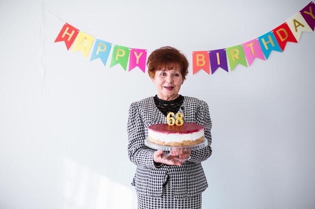 Starsza kobieta świętuje jej birthay w domu. Trzymając tort urodzinowy na białym tle