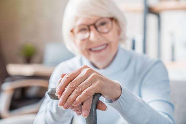 Starsza kobieta trzyma laskę siedząc na kanapie w domu z naciskiem na ręce