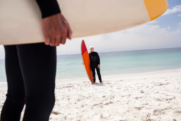 Zdjęcie starsza kobieta trzyma surfboard na plaży w kombinezonie