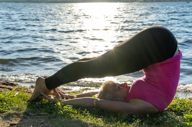 Starsza kobieta uprawia sport o świcie nad brzegiem jeziora Kobieta zarzuca nogi nad głowę Pozycja Halasana Joga dla zdrowia Elastyczne ciało Sporty na świeżym powietrzu