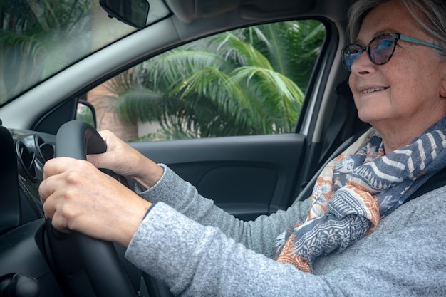 Starsza kobieta uśmiecha się po zaparkowaniu samochodu Kaukascy seniorzy z siwymi włosami i okularami Zielone rośliny za oknem samochodu