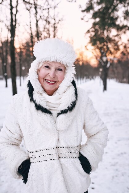 starsza kobieta w białym kapeluszu i futrze, ciesząc się zimą w lesie śniegu.