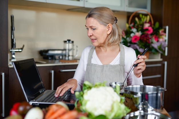 Starsza kobieta w kuchni przygotowuje jedzenie i zagląda do laptopa