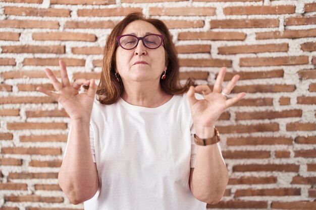Starsza kobieta w okularach stojąca nad murem z cegły zrelaksowana i uśmiechnięta z zamkniętymi oczami wykonująca gest medytacji palcami koncepcja jogi