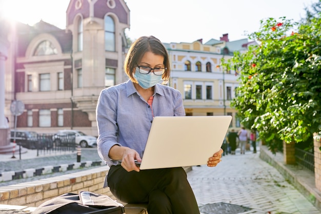 Starsza kobieta w twarzy medyczne maski ochronne z laptopem pracującym na zewnątrz. Tło miasta, biznes niezależny, koncepcja ochrony zdrowia