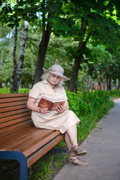 Starsza Kobieta Z Siwymi Włosami W Kapeluszu W Beżowej Sukience Siedząca Na ławce W Parku I Czytająca Książkę