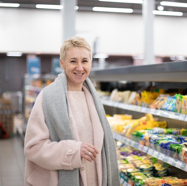 Starsza, ładna, dorosła, stylowa blondynka, uśmiechająca się do sklepu spożywczego, kupująca świeżą zdrową żywność w supermarkecie w sekcji zimnych sklepów spożywczych, patrząca na kamerę