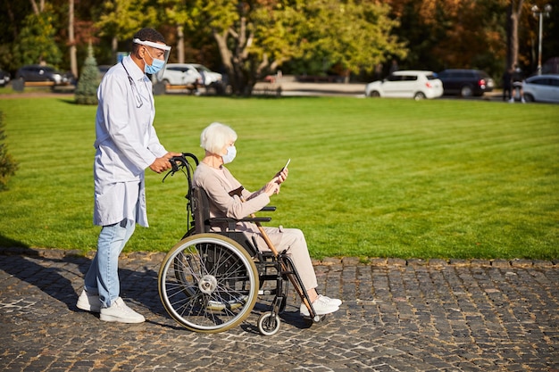Starsza Pani Siedzi Ze Smartfonem W Dłoniach, Podczas Gdy Pracownik Medyczny W Tarczy Ochronnej Pcha Jej Wózek Inwalidzki