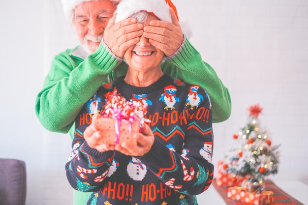 Starsza para w ciepłych ubraniach i santa hat mężczyzna zasłaniający oczy starej żony prezentem niespodzianką dla niej Kochająca stara romantyczna heteroseksualna para świętująca wspólnie świąteczny festiwalxA