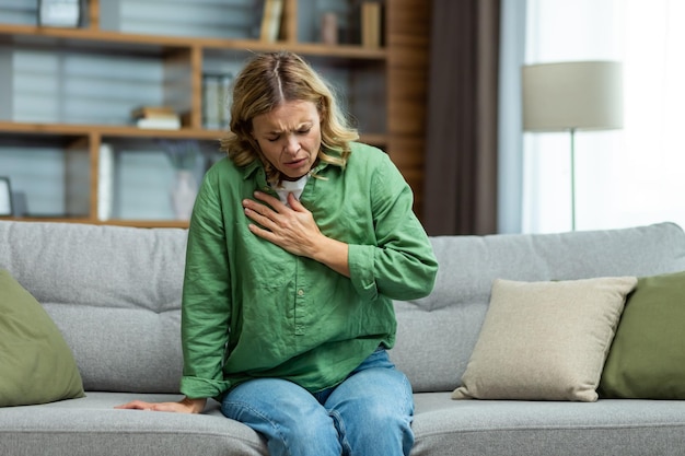 Starsza piękna kobieta siedząca chora na kanapie w domu trzyma jego klatkę piersiową, ma alergię na astmę