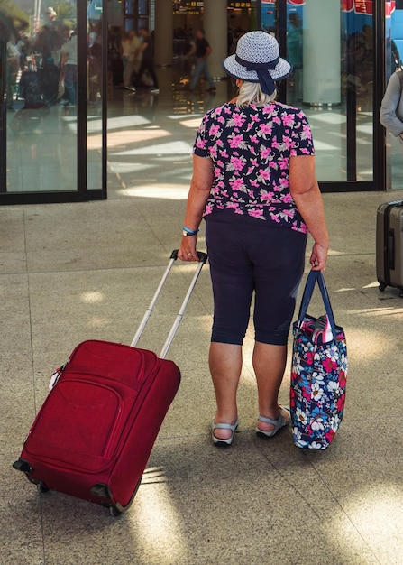 Starsza starsza kobieta w letnich ubraniach wakacyjnych ciągnąca swój bagaż wchodząc do terminalu lotniska, widok od tyłu