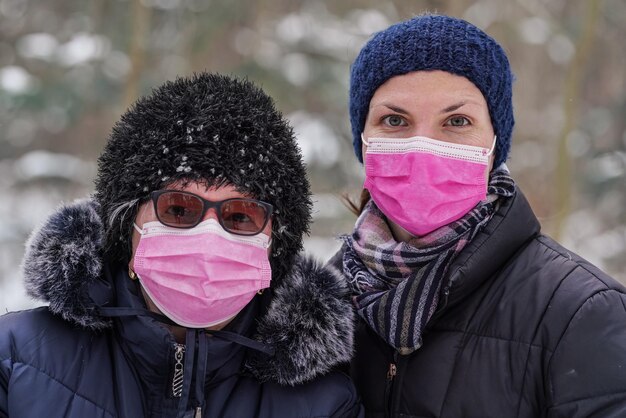 Starsza starsza kobieta z młodszą córką w ciepłych zimowych ubraniach, oboje w różowej jednorazowej masce na twarz z wirusem