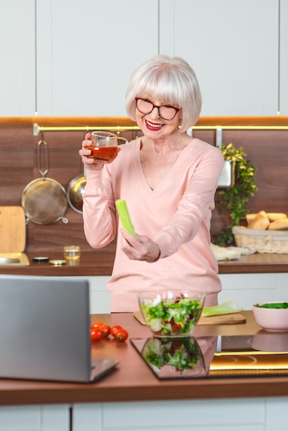 starsza, wesoła kobieta, blogerka wideo pokazująca seler i sok pomidorowy w swojej kuchni