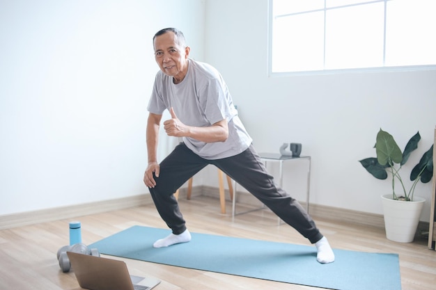 Zdjęcie starszy azjat ćwiczy za pomocą laptopa, ma online zajęcia fitness z domu i pokazuje kciuki.