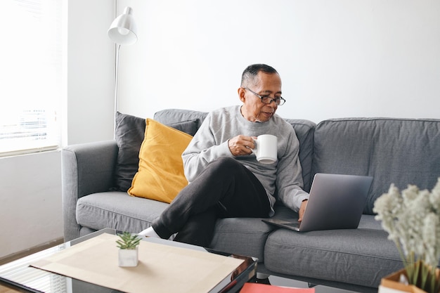Zdjęcie starszy azjat siedzący na kanapie i używający laptopa, trzymając filiżankę kawy w domu