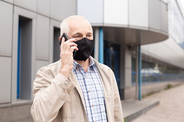 Starszy Biznesmen Noszenie Maski Za Pomocą Rozmowy Ze Smartfonem W Mieście, W Pobliżu Budynków Biurowych