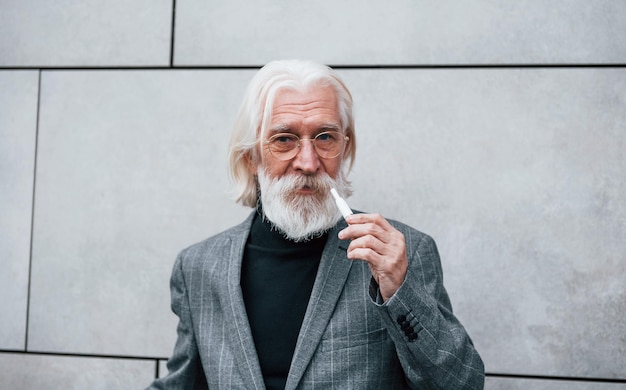 Starszy biznesmen w formalnych ubraniach z siwymi włosami i brodą jest na zewnątrz, pali elektroniczny papieros
