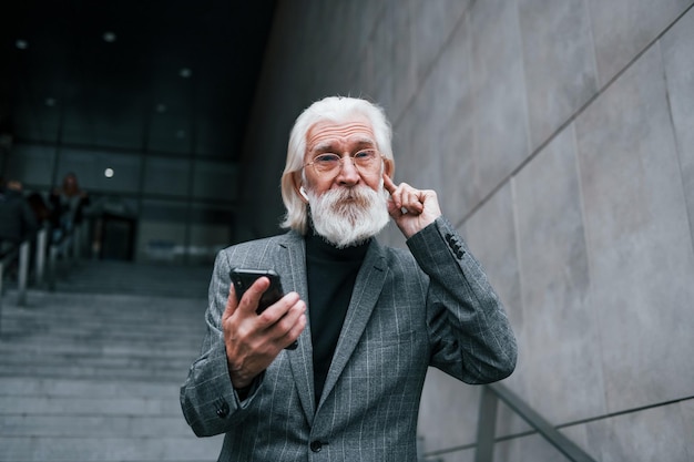 Starszy biznesmen w formalnych ubraniach z siwymi włosami i brodą jest na zewnątrz, próbując słuchawek bezprzewodowych z telefonem