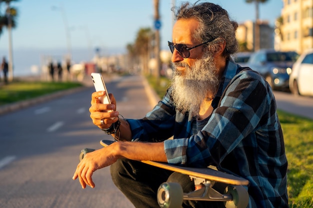Starszy broda mężczyzna z deskorolką, siedząc na ulicy, ciesząc się zachodem słońca