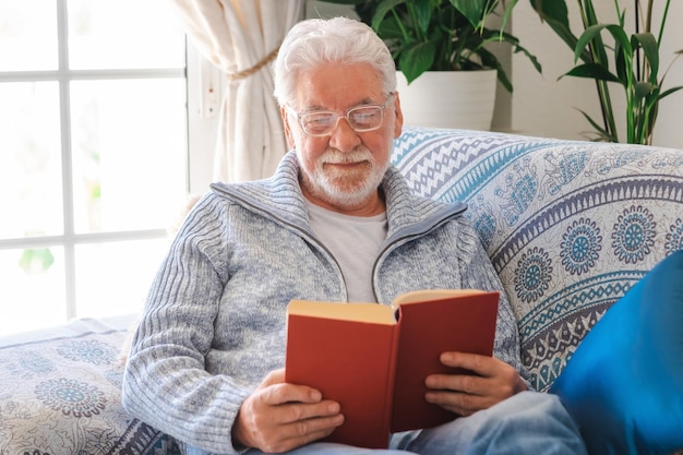 Starszy brodaty mężczyzna relaksuje się na kanapie w domu czytając książkę Senior w okularach i jasnoniebieskim swetrze