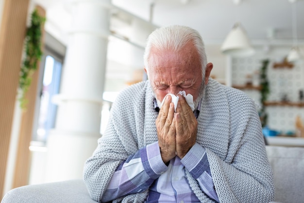 Starszy chory brodaty mężczyzna kicha do serwetki w domu na szarej kanapie z białym kocem Ochrona przed chorobami wirus koronawirusa choroba grypy opatrunek oddechowy
