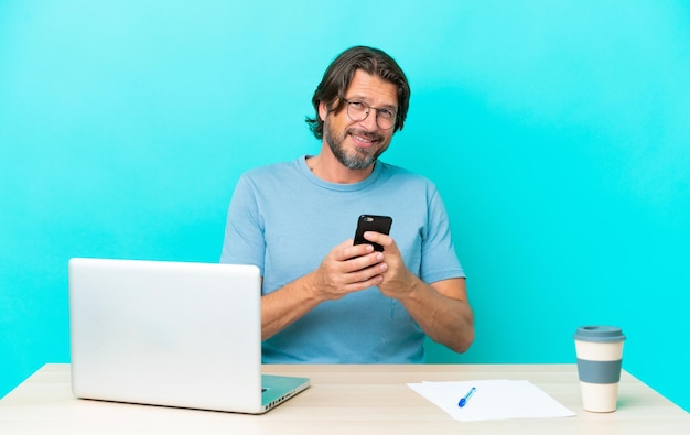 Starszy holenderski mężczyzna przy stole z laptopem na białym tle na niebieskim tle wysyła wiadomość za pomocą telefonu komórkowego