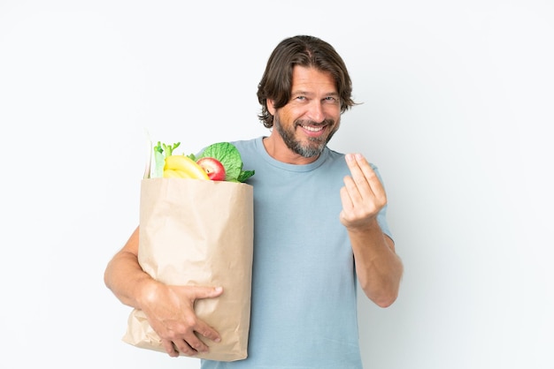 Starszy holenderski mężczyzna trzymający torbę na zakupy spożywcze na białym tle robiący gest pieniędzy